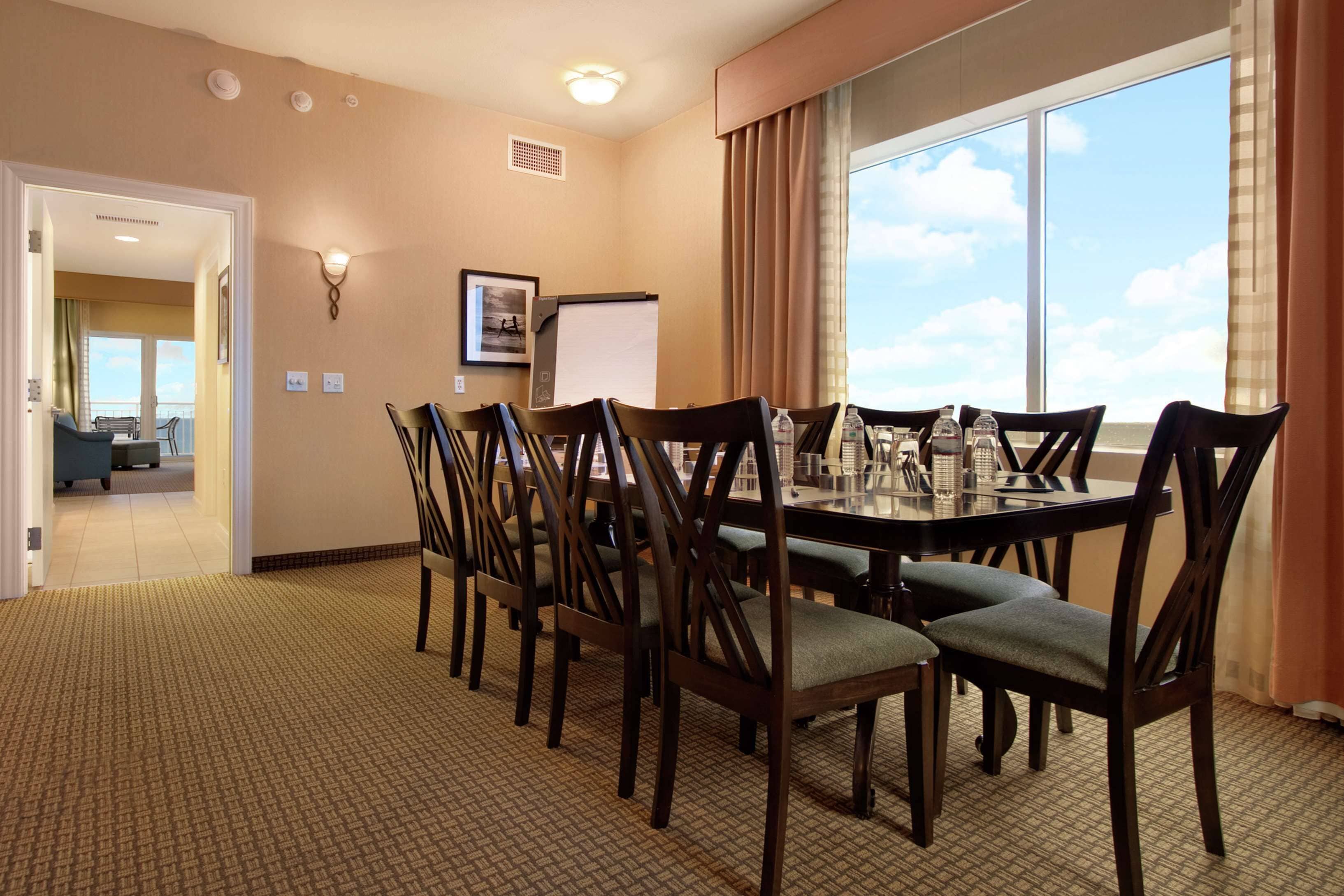 A conference room with a large table, chairs, a projector screen, and a view of the sky through a large window.