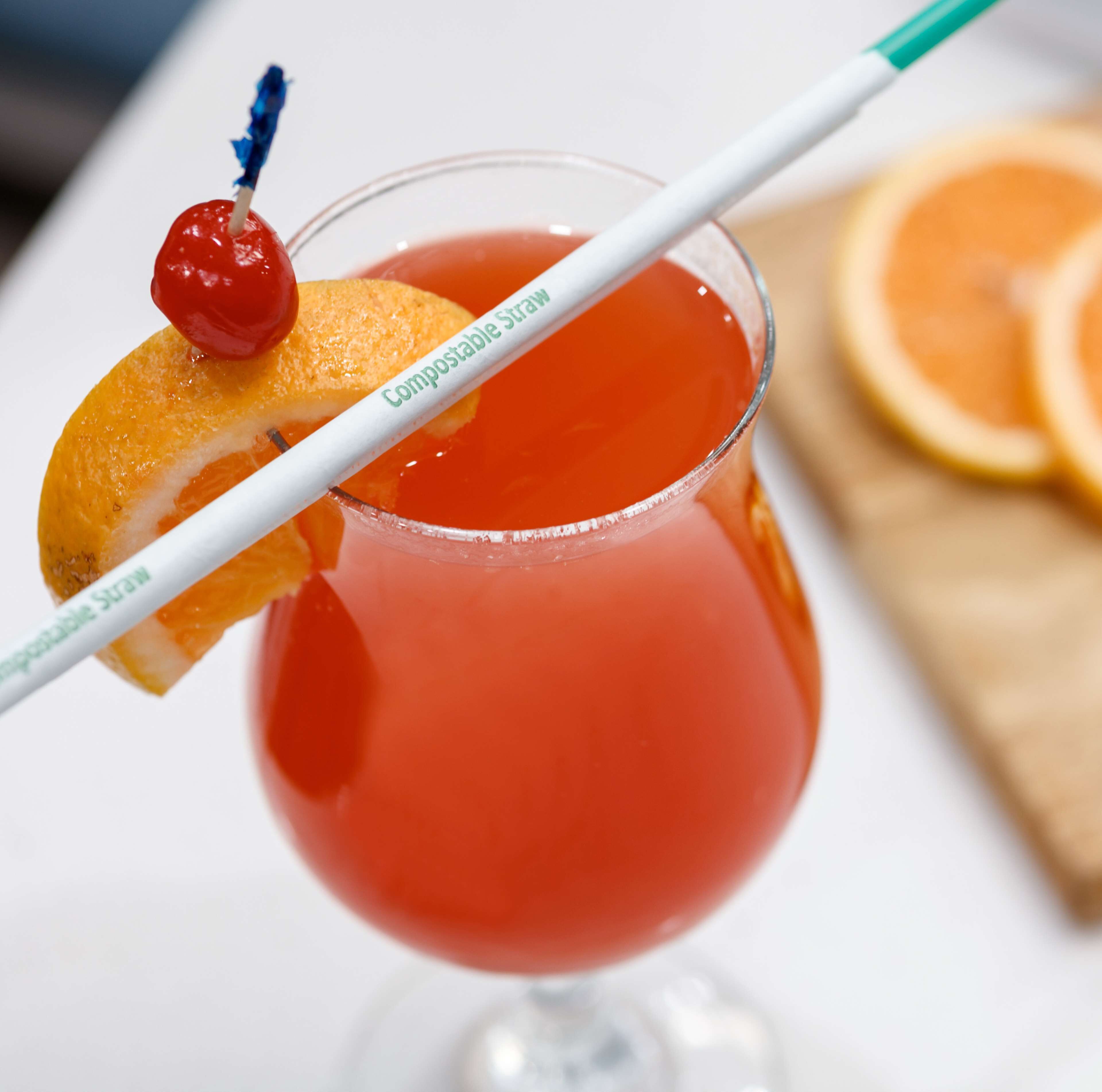 A cocktail with a cherry and orange slice garnish, served in a glass with a straw labeled "Compostable Straw."