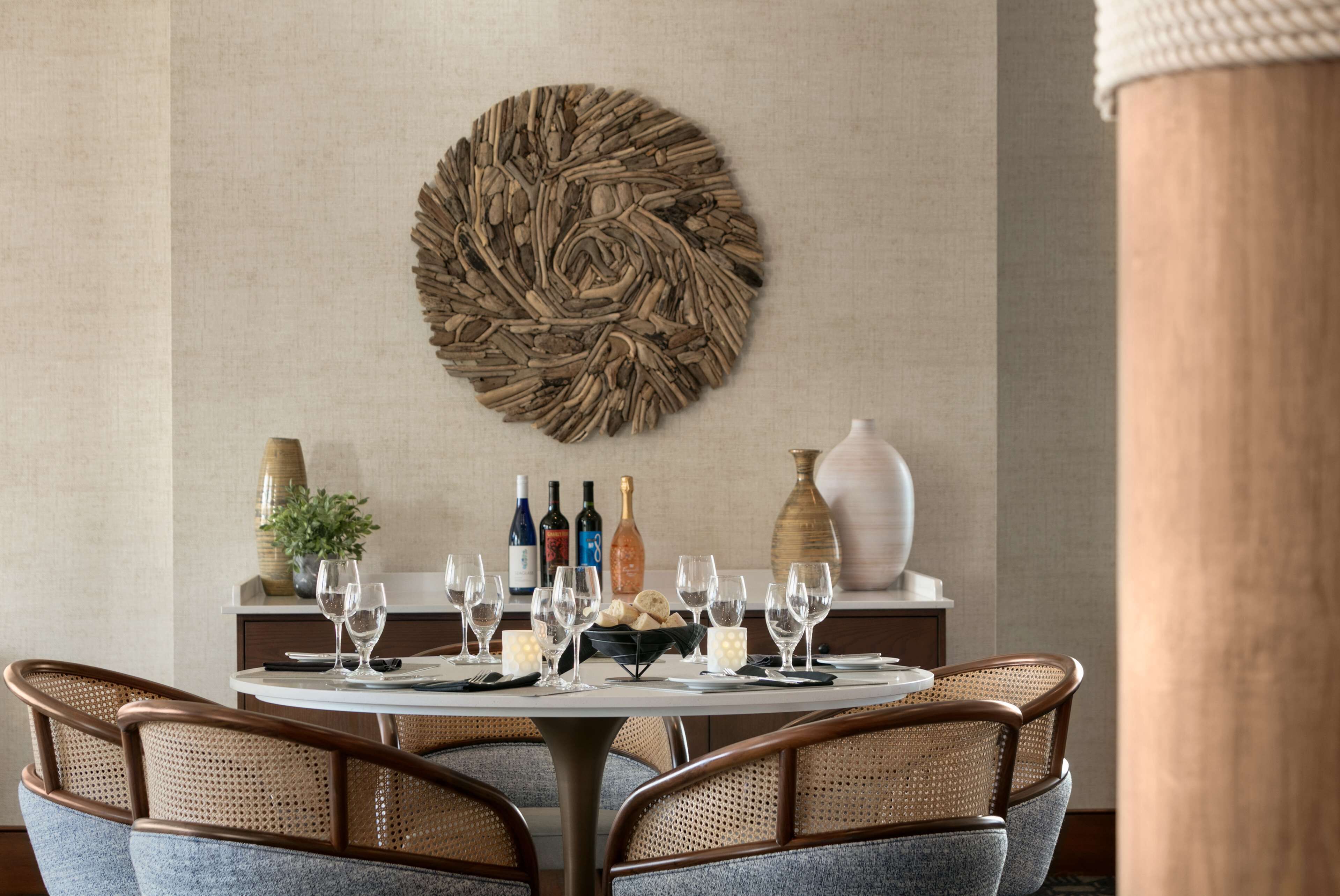 A dining area with a round table set for six, surrounded by wicker chairs. A decorative wall piece and a vase are visible on the wall behind the table.