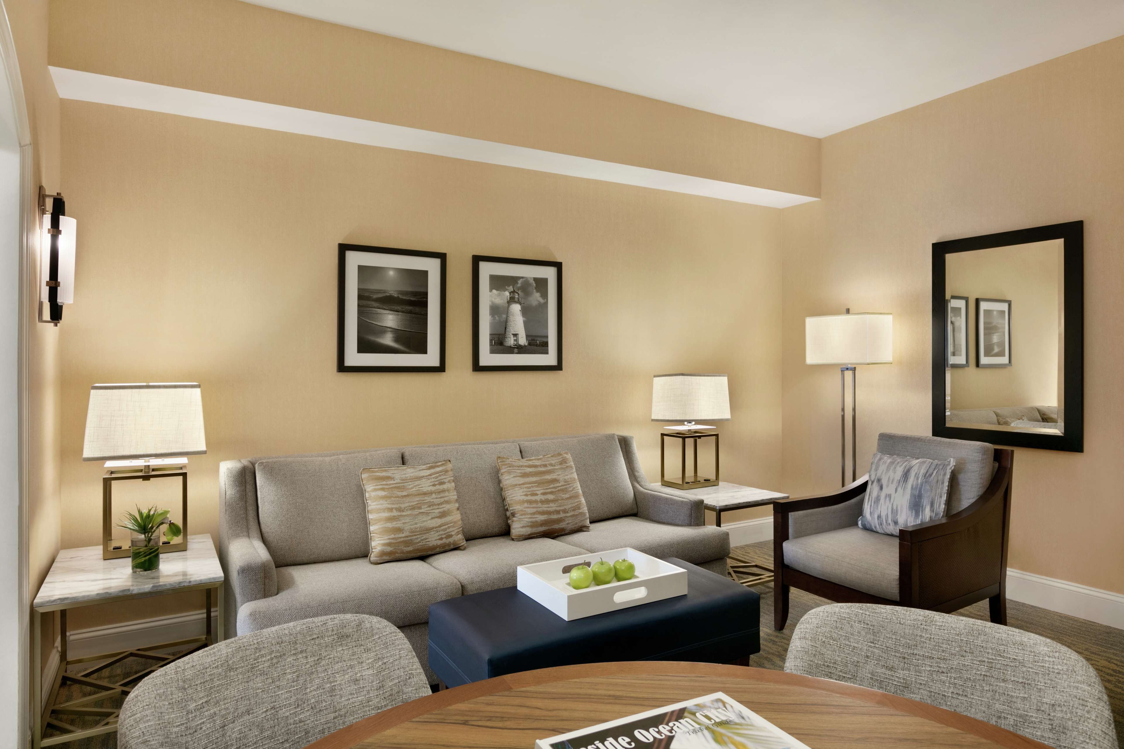 A modern hotel room with a grey sofa, a small round table with a magazine, a side table with a lamp, and a chair with a cushion.