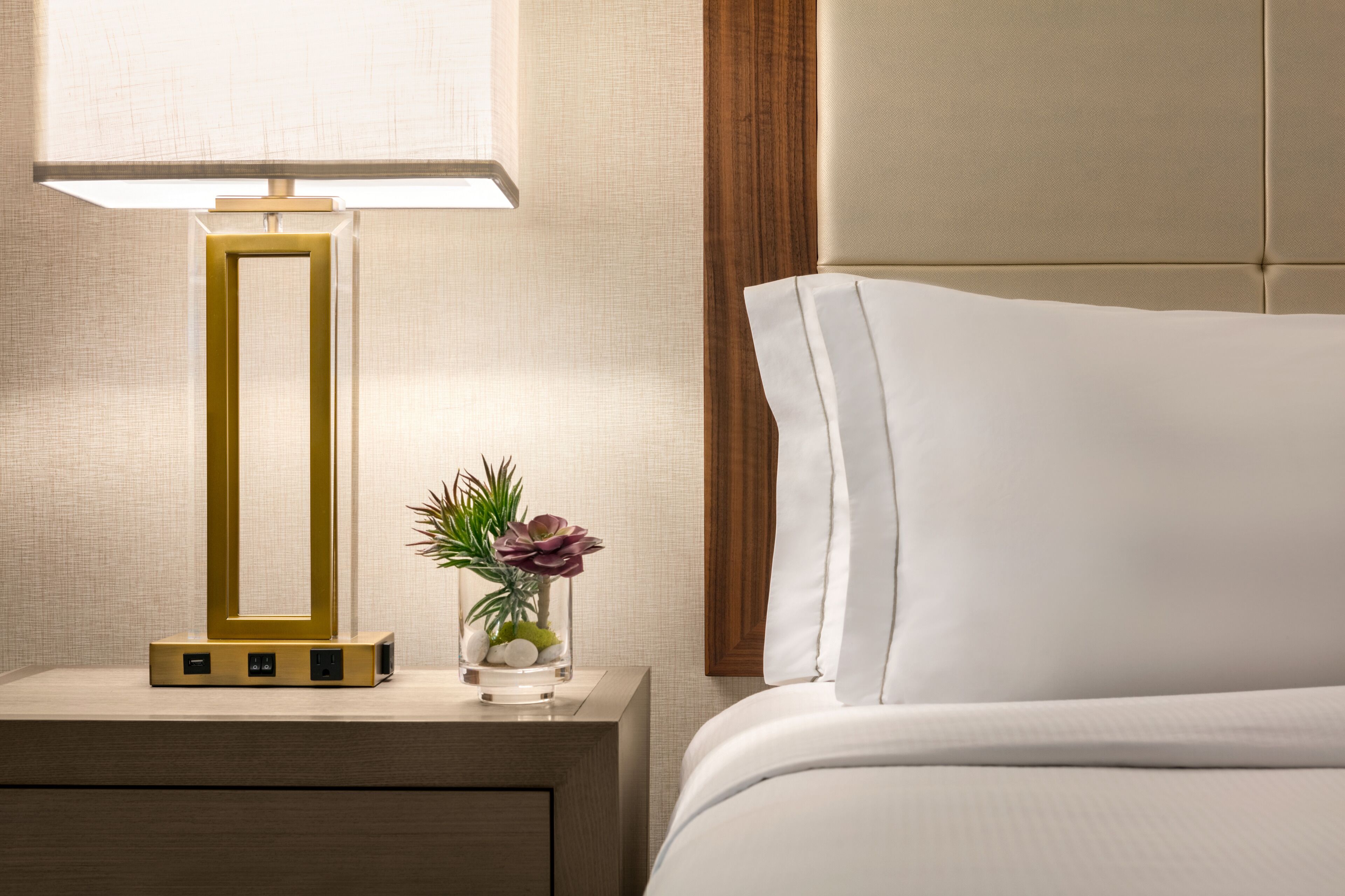 A neatly made bed with a white pillow and a headboard, a bedside table with a lamp, and a small vase with flowers.