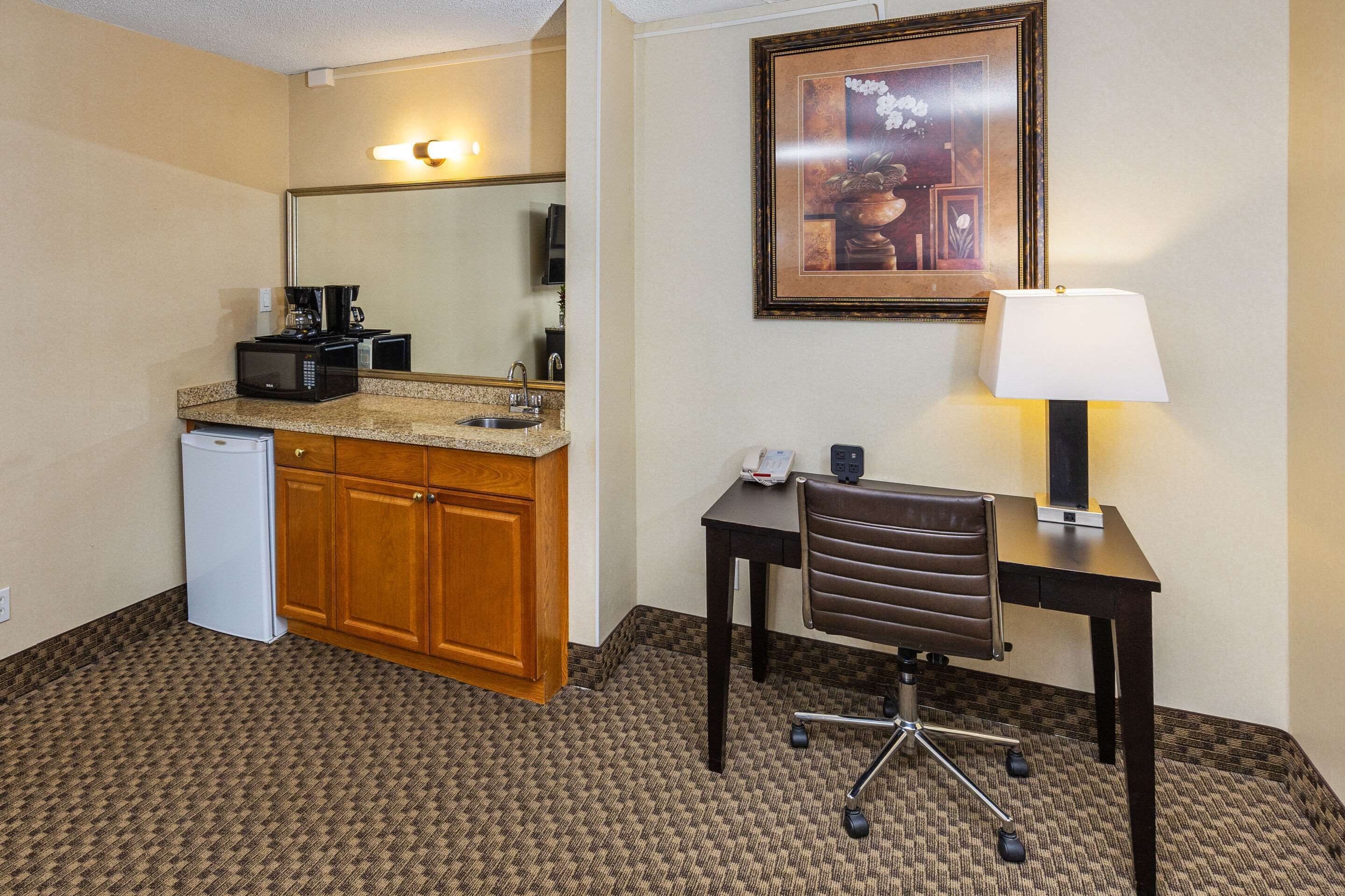 A hotel room with a kitchenette, a desk with a chair, a lamp, and a framed picture on the wall.