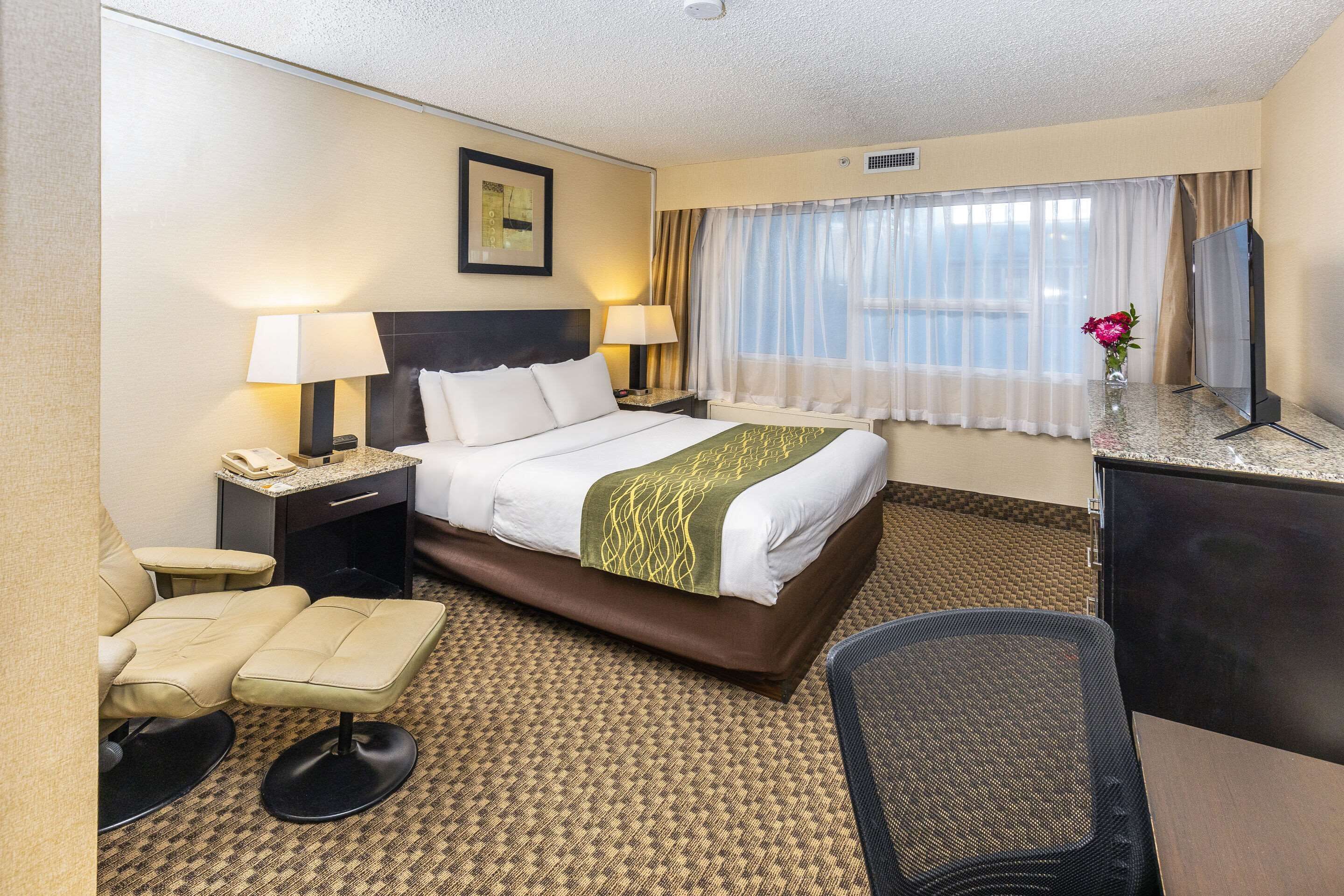 Una habitación de hotel con una cama, dos lámparas de noche, un escritorio con teléfono, una silla, un televisor y una ventana con cortinas.