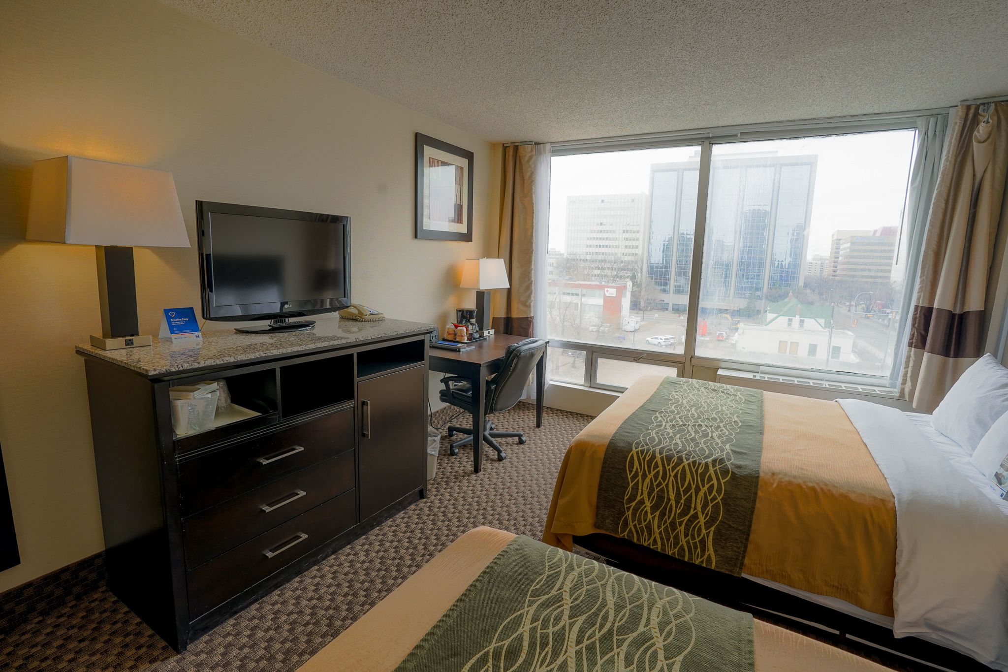 Habitación de hotel con cama, escritorio, televisor y un amplio ventanal con vistas a la ciudad.