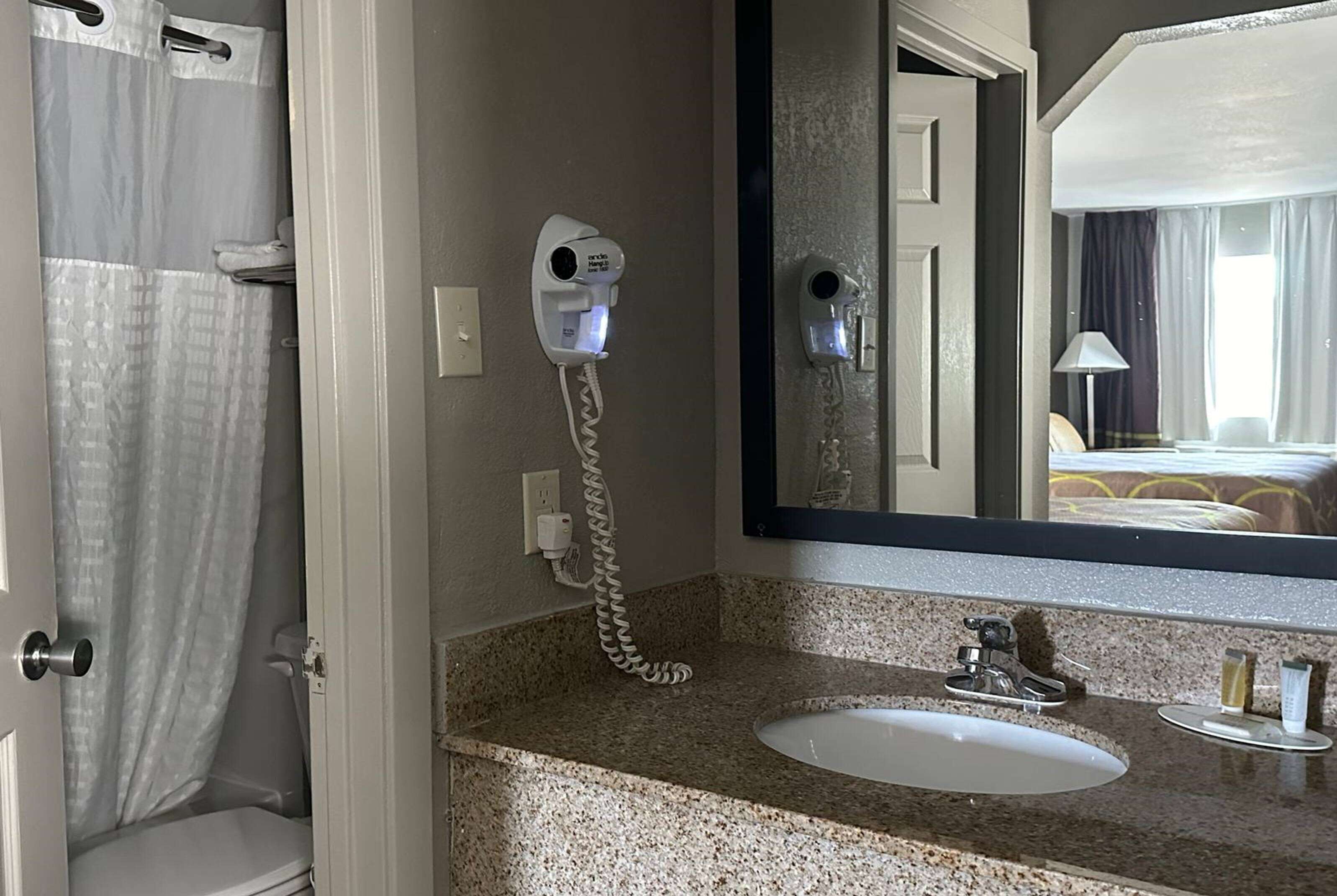 A bathroom with a granite countertop, a white sink, a hairdryer, and a mirror reflecting a bed and curtains.