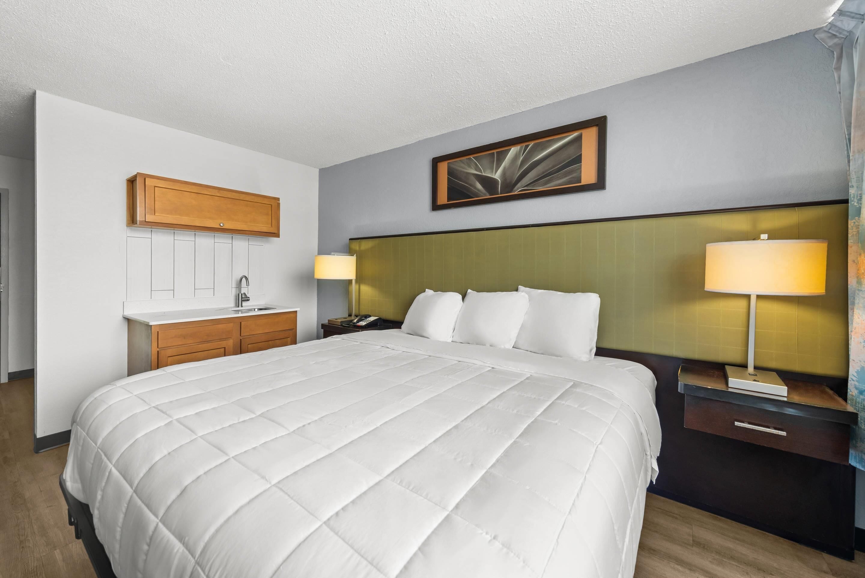 A hotel room with a large bed, a bedside table with a lamp, a small desk with a lamp, a wooden cabinet, and a framed picture on the wall.