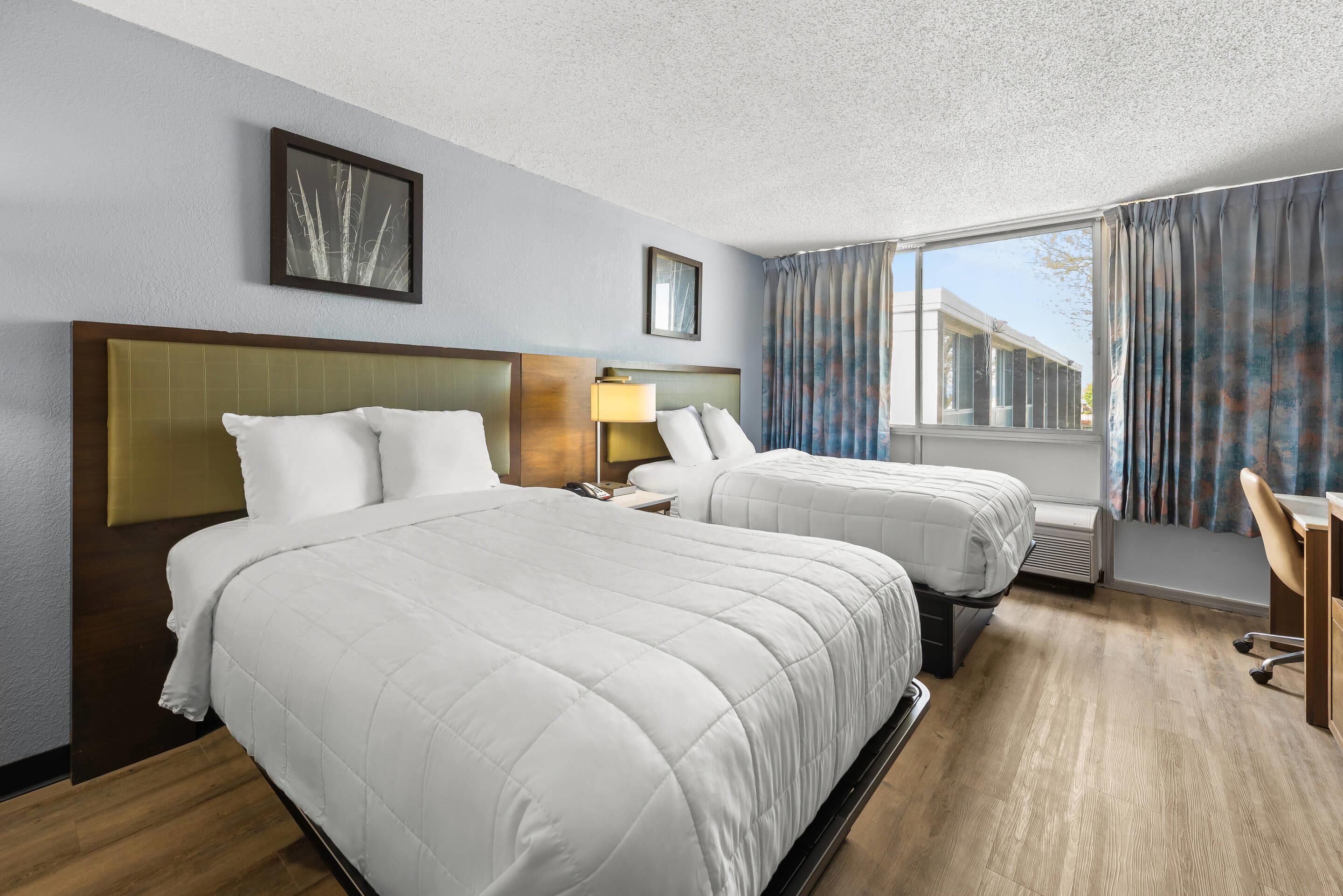 A hotel room with two beds, a desk, a chair, a lamp, and a window with curtains.