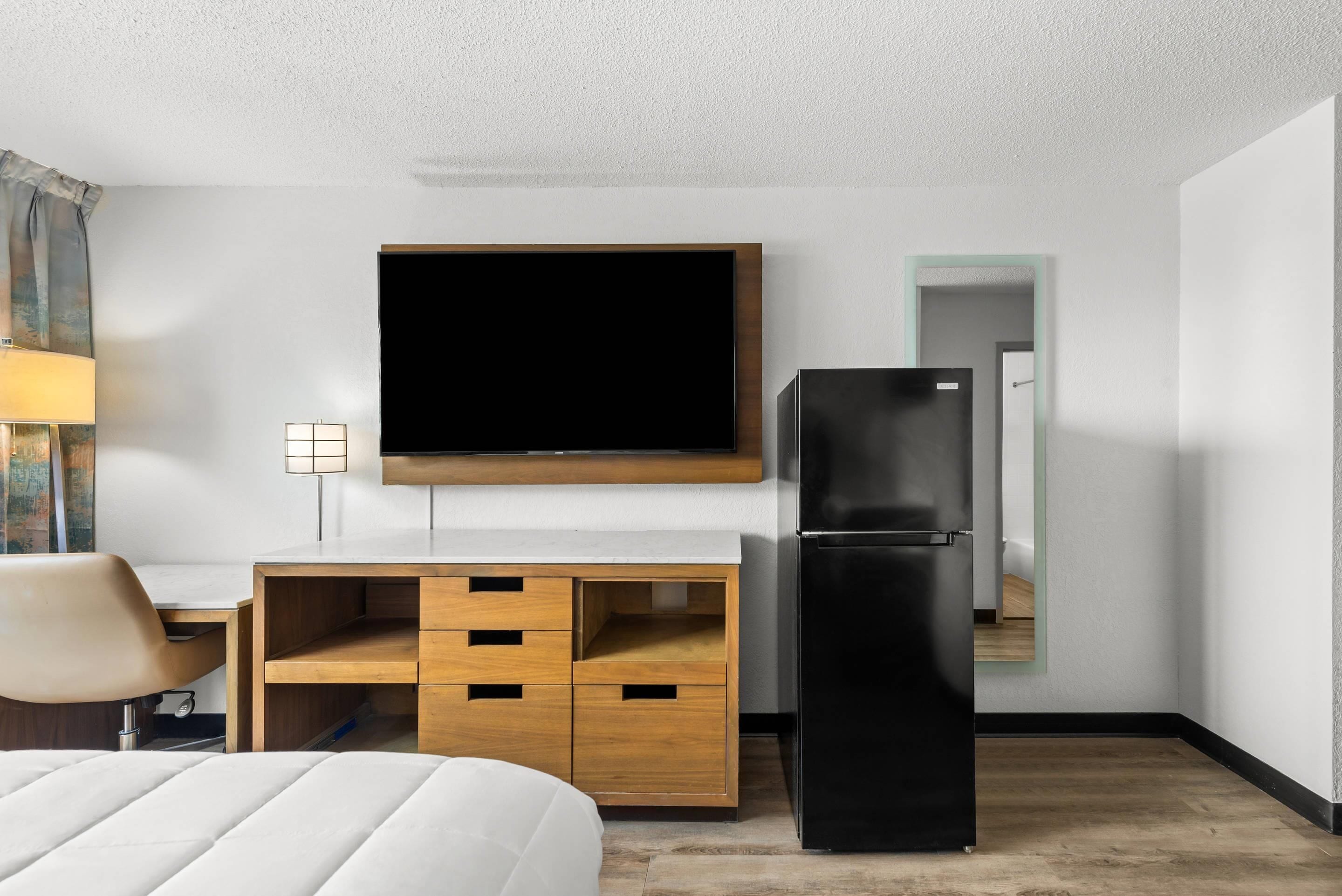 A hotel room with a bed, a desk with a chair, a flat-screen TV, a black refrigerator, and a wooden cabinet.