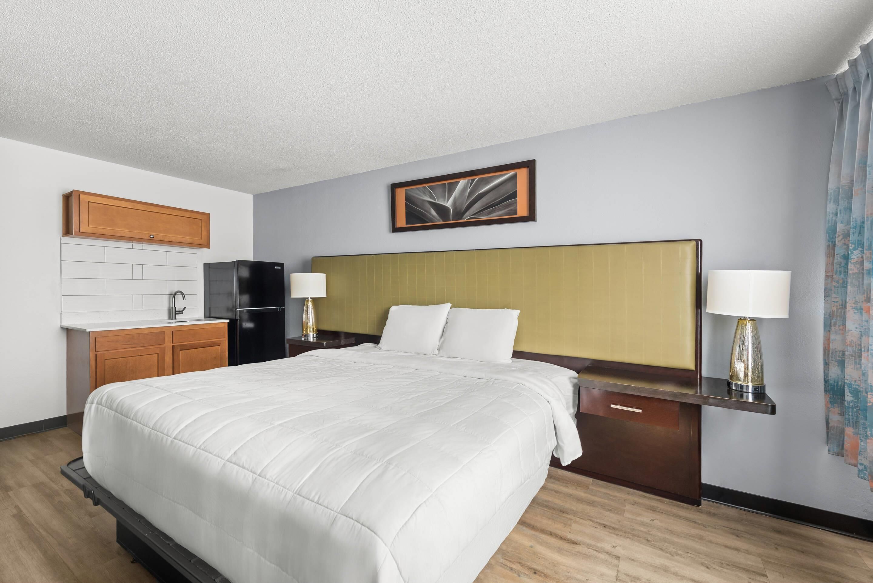 Habitación de hotel con una cama grande, cabecera de madera, kitchenette pequeña y un cuadro enmarcado en la pared.
