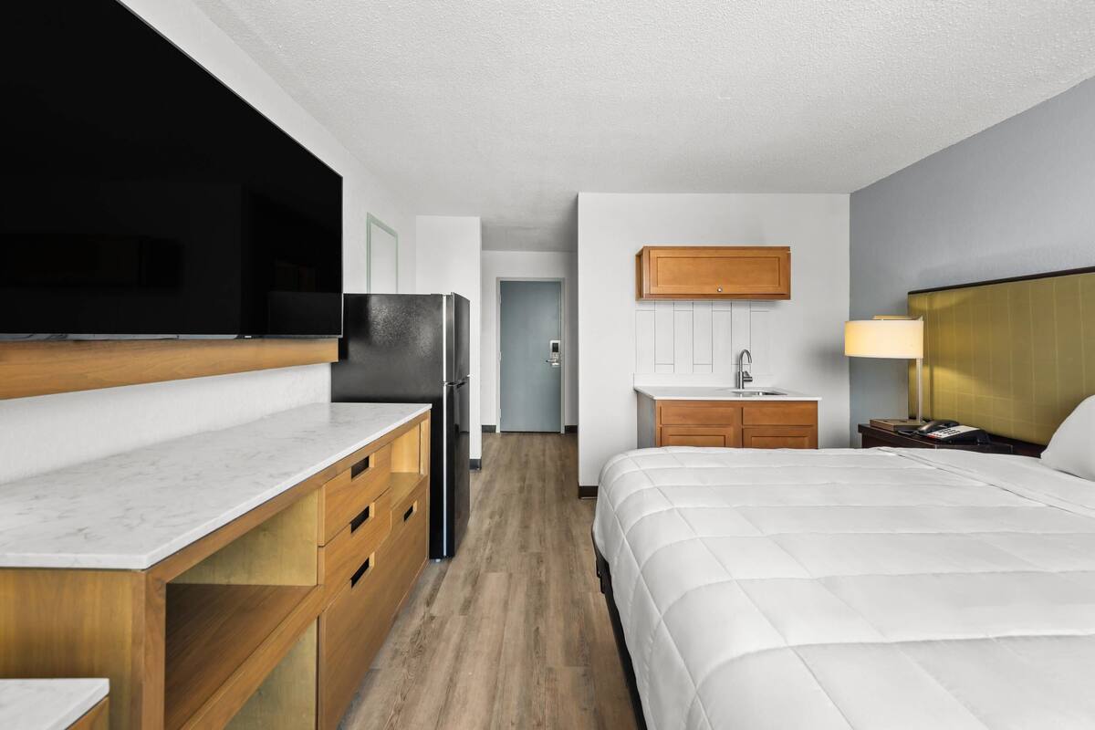 A modern hotel room with a wooden desk, a bed, a television, a sink, and a nightstand.