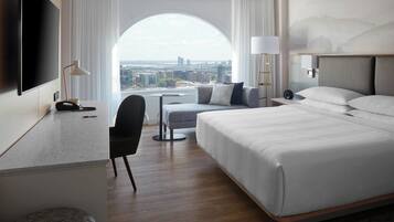 A hotel room with a large bed, a desk with a chair, a TV, and a view of the city through a large window.
