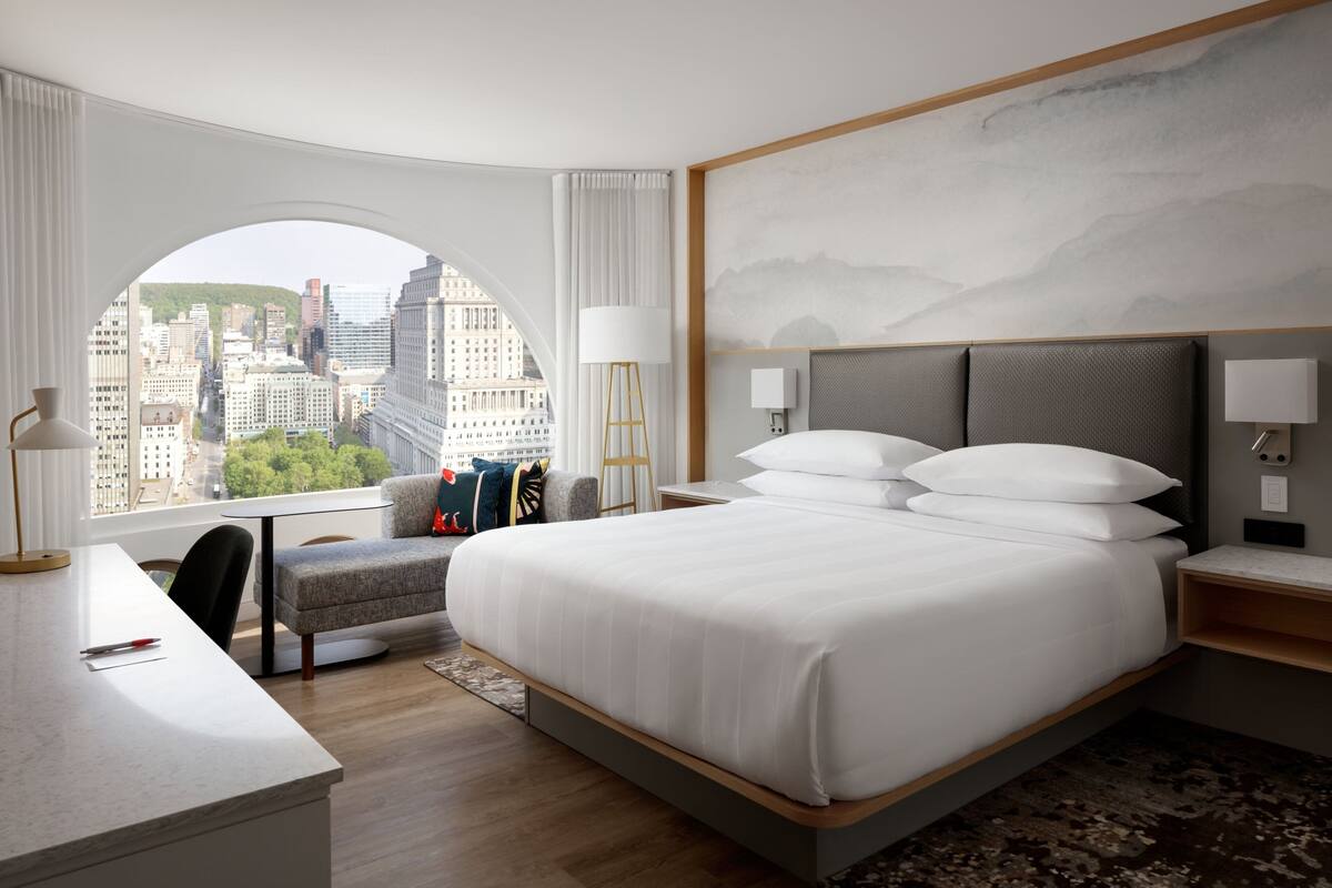 A modern hotel room with a large bed, a desk, and a view of a cityscape through a large window.