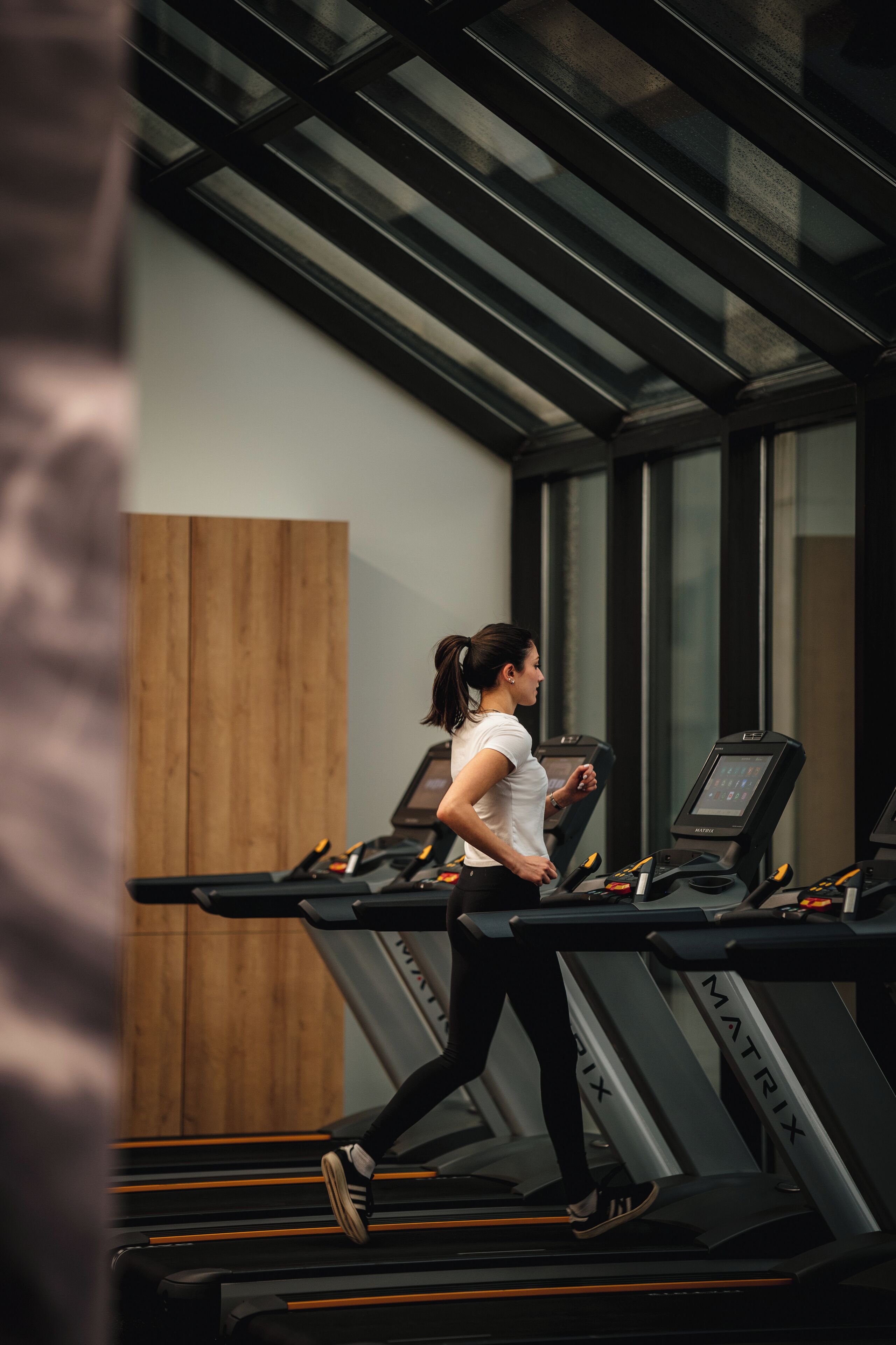 Una mujer corriendo en una caminadora en un gimnasio con paneles de madera y ventanas amplias.
