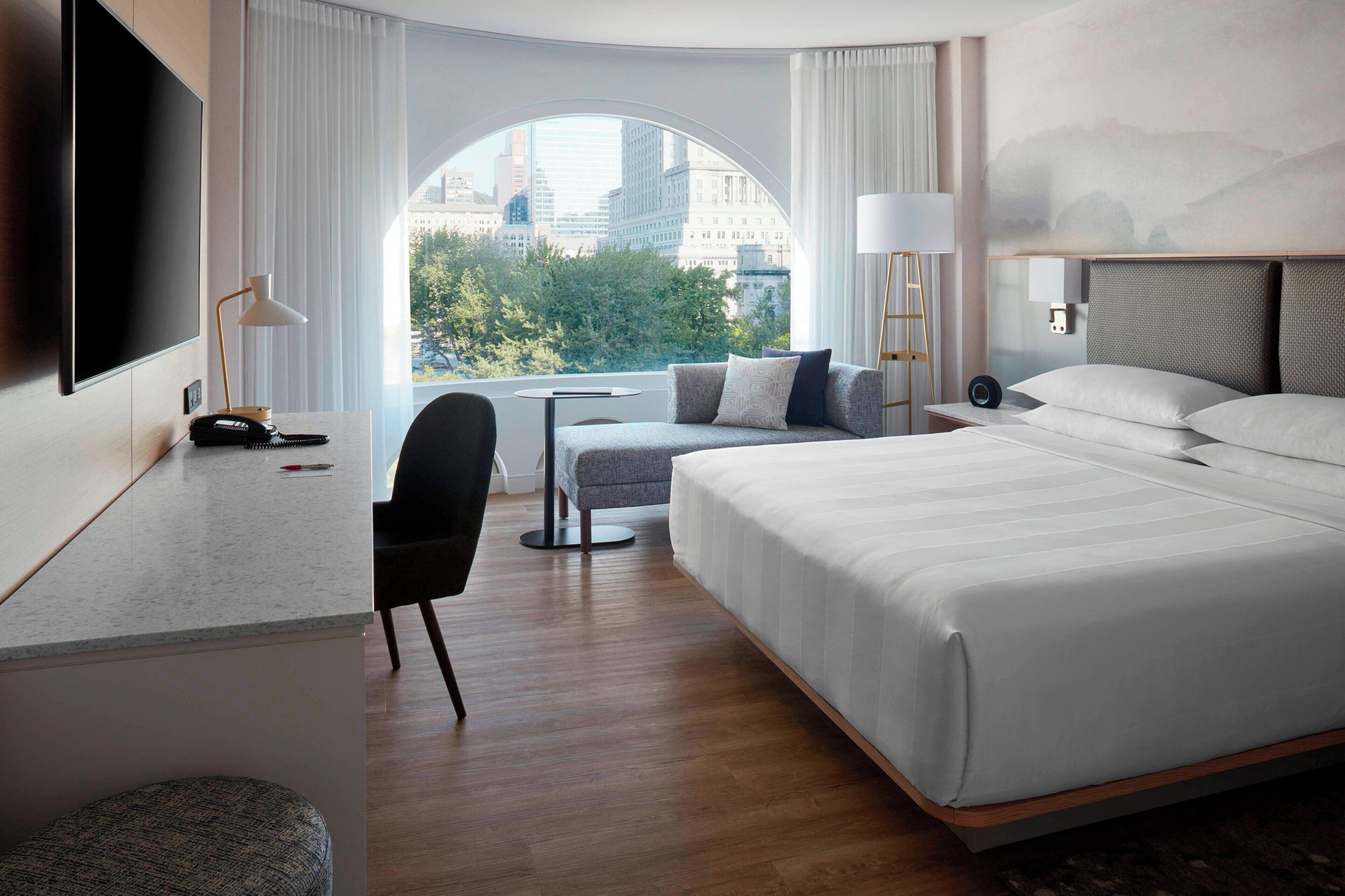 Una habitación de hotel moderna con una cama grande, un escritorio con silla, una mesa redonda y un ventanal con vistas a la ciudad.