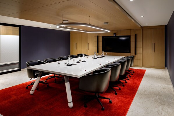 Un salón de conferencias con una larga mesa, sillas negras, alfombra roja, armarios de madera y un televisor de pantalla plana grande.