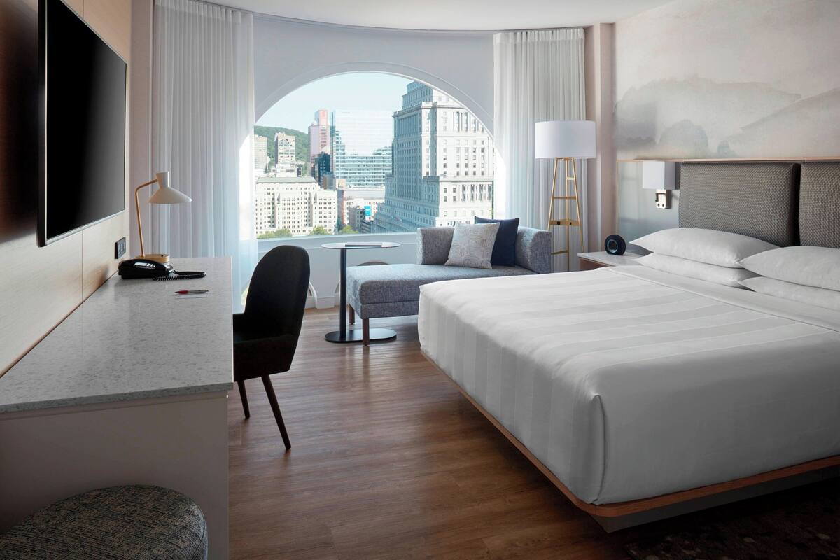 A modern hotel room with a large bed, a desk, a chair, and a view of the cityscape through a large window.