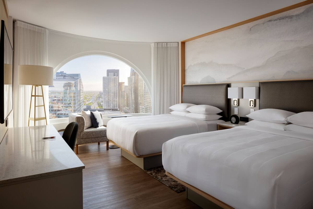 A modern hotel room with two beds, a desk, and a large window offering a city view.