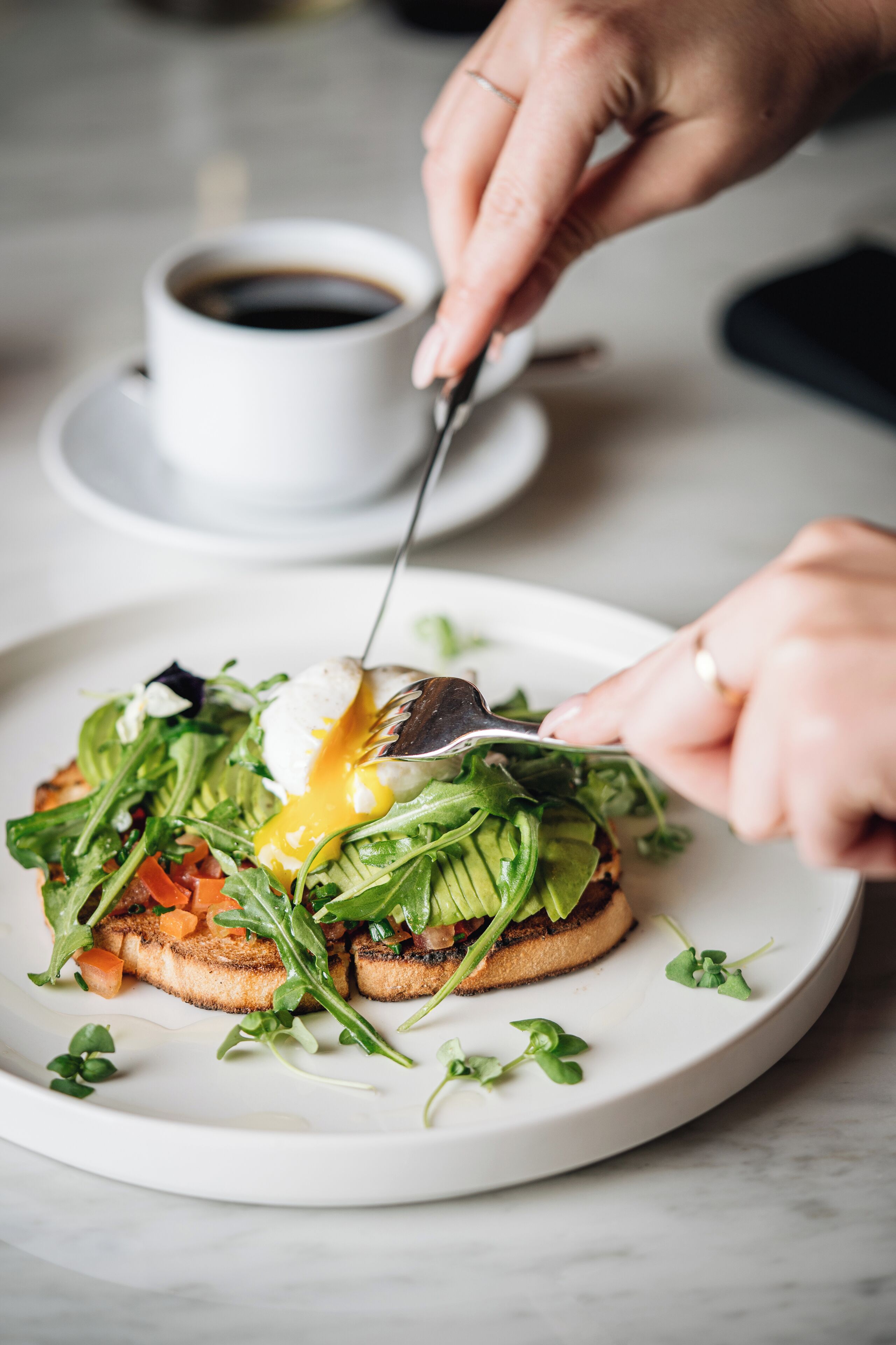 Une assiette de nourriture surmontée d’un œuf poché, agrémentée de verdure et de légumes, et une tasse de café en arrière-plan.