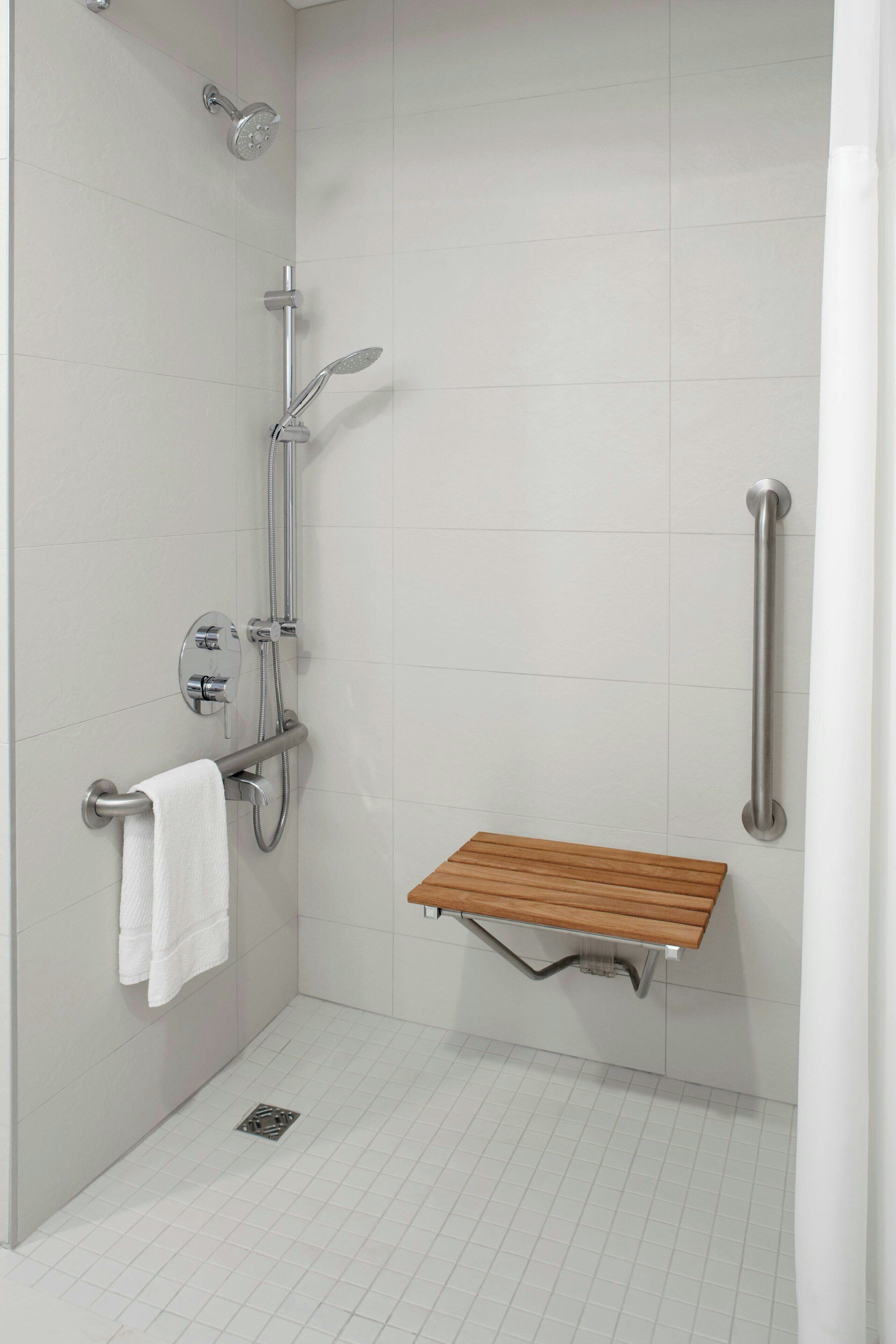 Ducha moderna con cabezal de ducha manual, cabezal de ducha tipo lluvia manual y grifo empotrado. Hay un pasamanos a la derecha y un banco de madera en la parte inferior.
