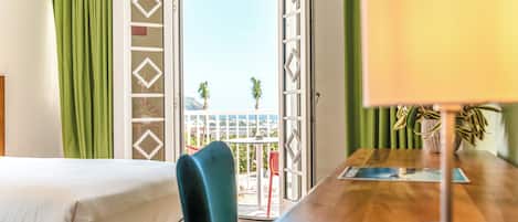 A hotel room with a bed, a wooden table with a lamp, a chair, and a view through a glass door to the outside.