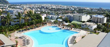 Un resort con una gran piscina rodeada de sillones de descanso, palmeras y vistas al océano y al paisaje urbano.