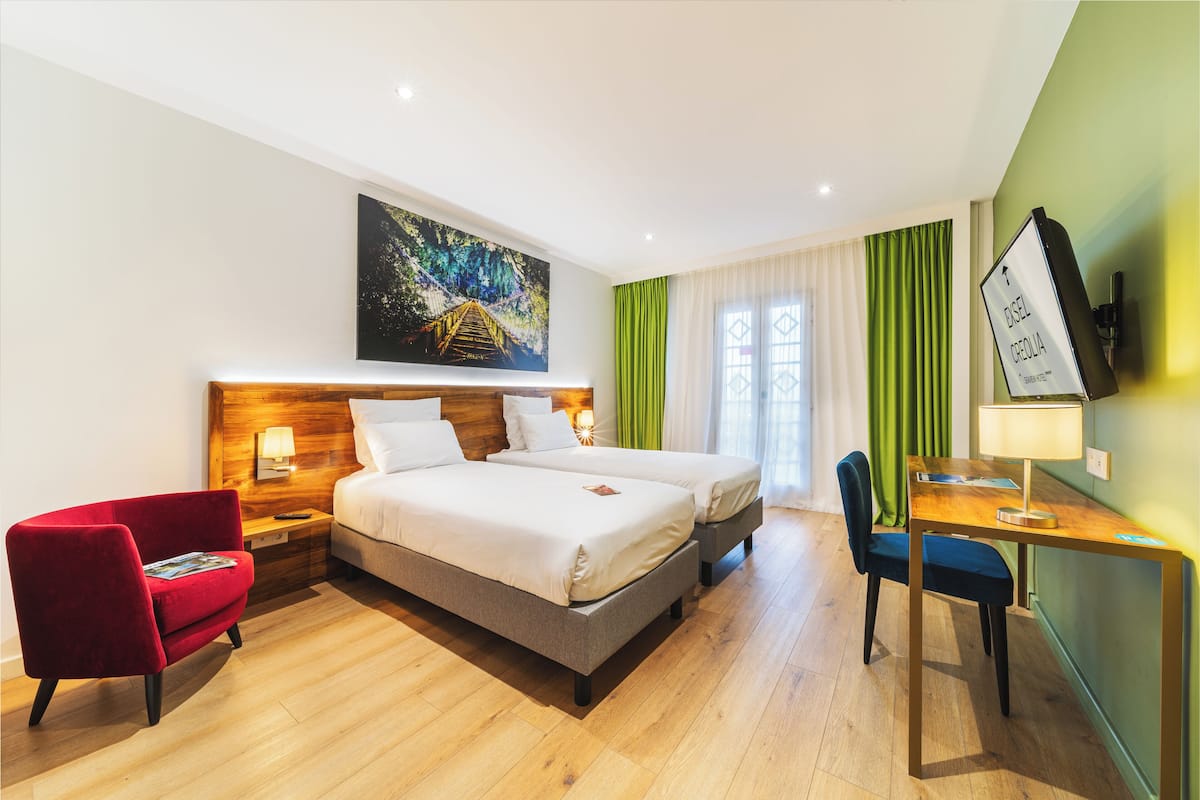 A hotel room with a bed, a desk, a chair, a nightstand, a lamp, and a painting on the wall.