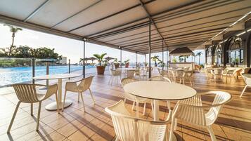 Un patio al aire libre con mesas redondas y sillas blancas, con vista a una piscina y palmeras.