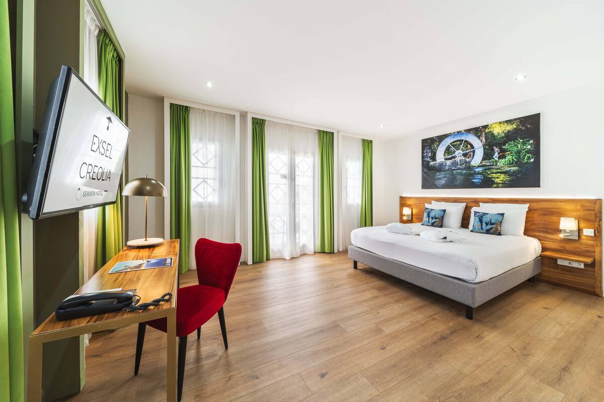 A hotel room with a bed, desk, chair, TV, and artwork on the wall.