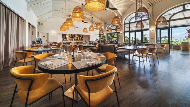 A spacious dining area with round tables, yellow chairs, and large windows offering views of the outdoors. - Hôtel Exsel Créolia  (Saint-Denis)
