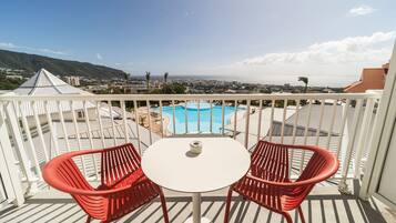 Un balcon avec une balustrade blanche, deux chaises rouges, une petite table blanche et une vue sur une piscine et le paysage urbain au loin.