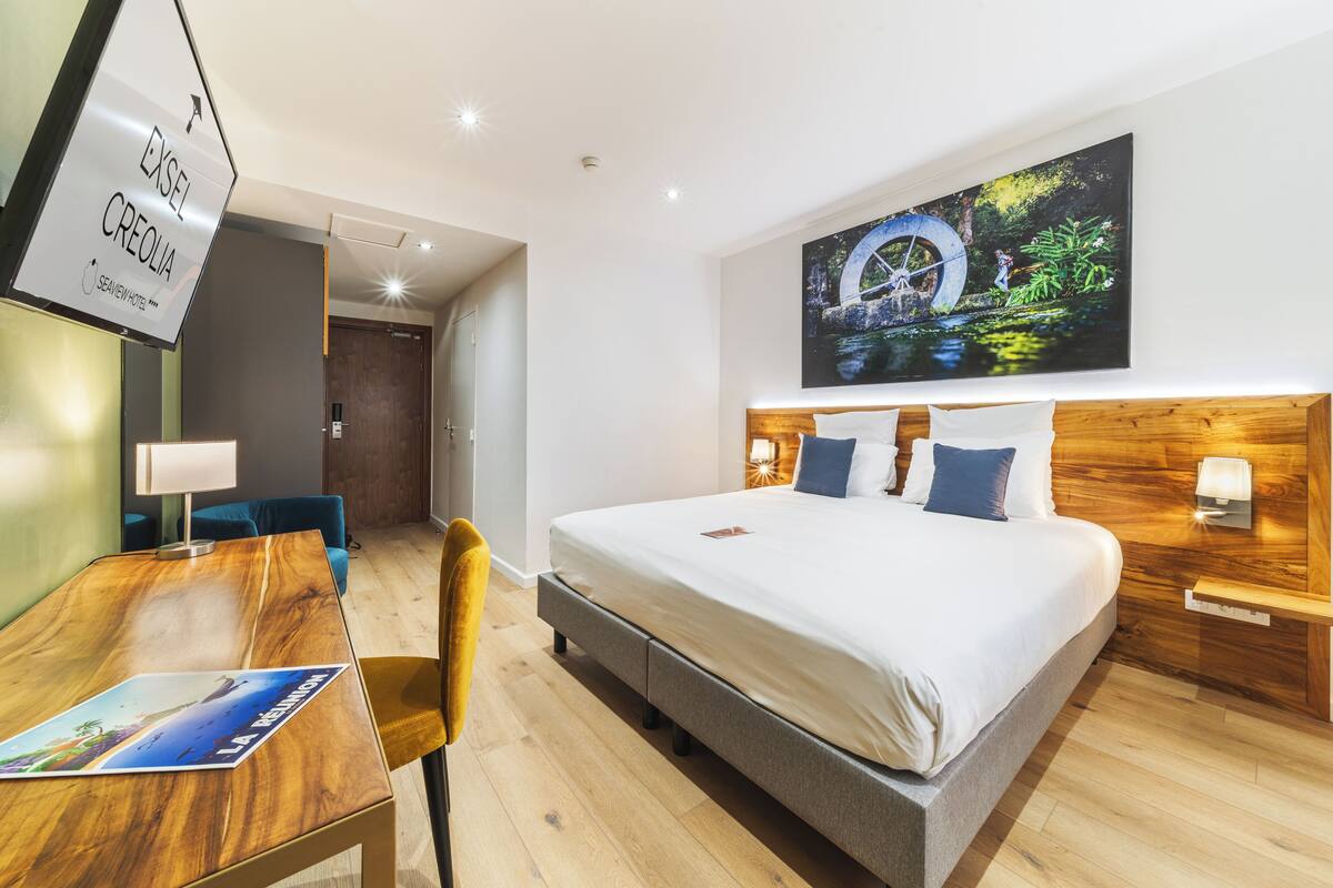 A hotel room with a bed, a desk, a chair, a wooden headboard, a nightstand, and a wall-mounted artwork.