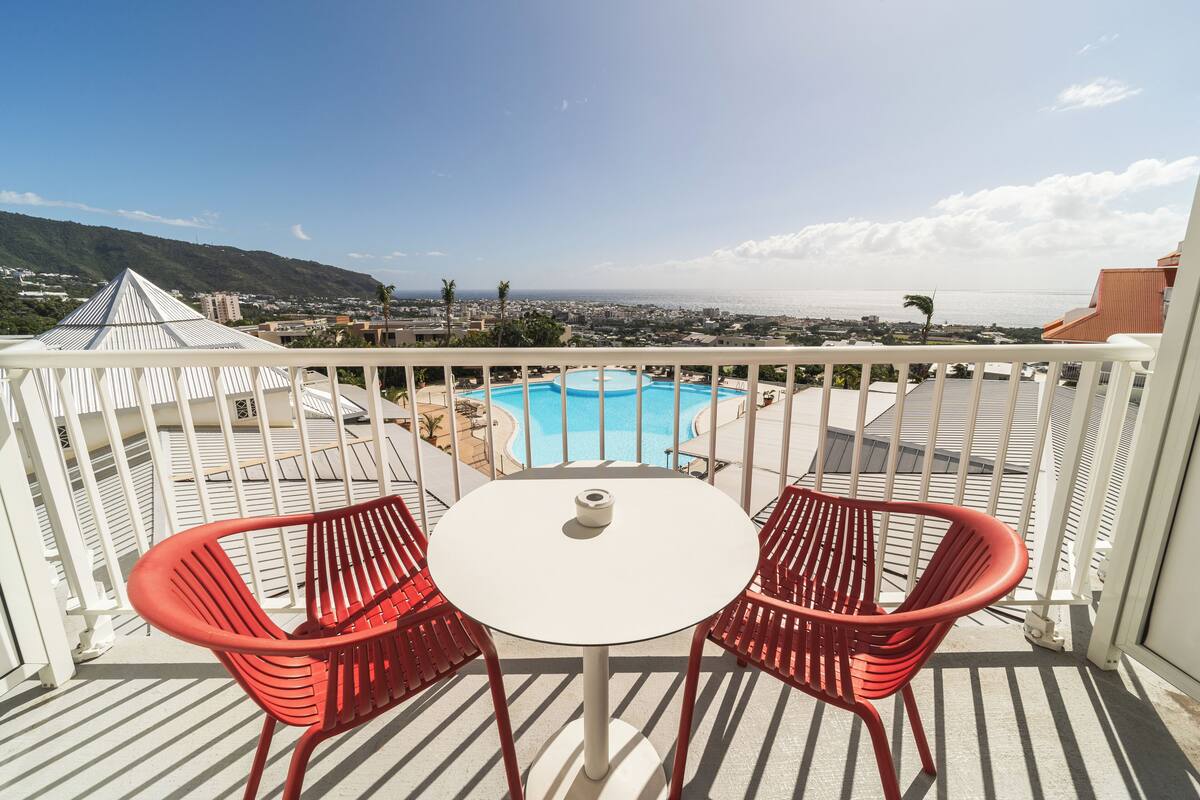 A balcony with two red chairs and a small table, overlooking a pool and a coastal cityscape.