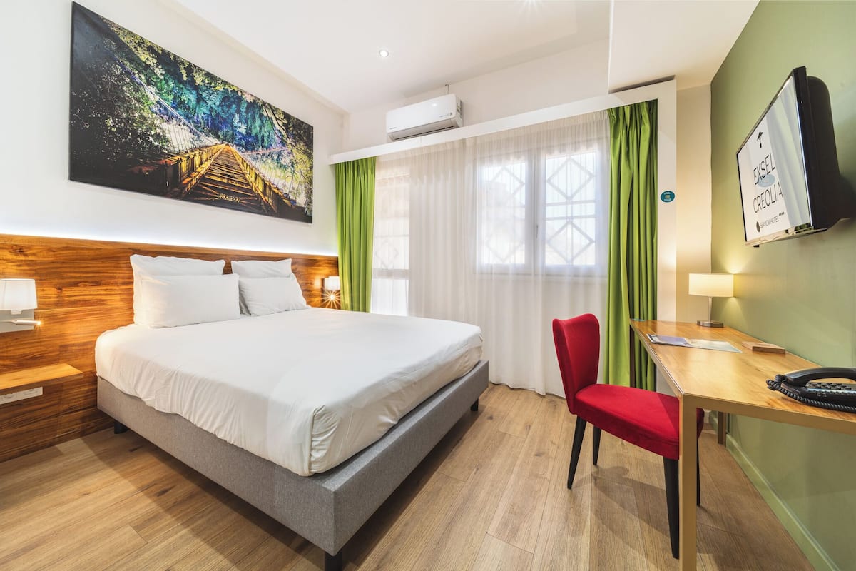 A hotel room with a bed, a desk, a chair, a TV, and a painting on the wall.
