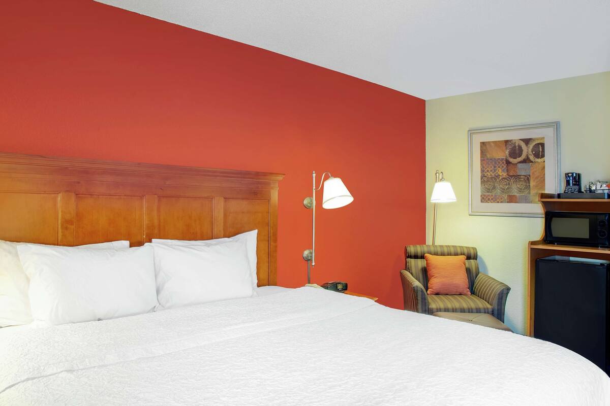 A hotel room with a large bed, a wooden headboard, a small armchair, a desk with a lamp, a microwave, and a refrigerator.