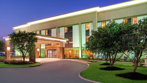 A modern hotel entrance with a curved driveway, a welcoming sign, and well-maintained landscaping.