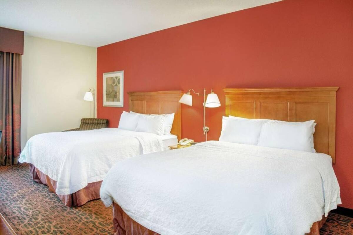 Two double beds with white linens, a wooden headboard, a bedside table with a lamp, and a patterned carpet.
