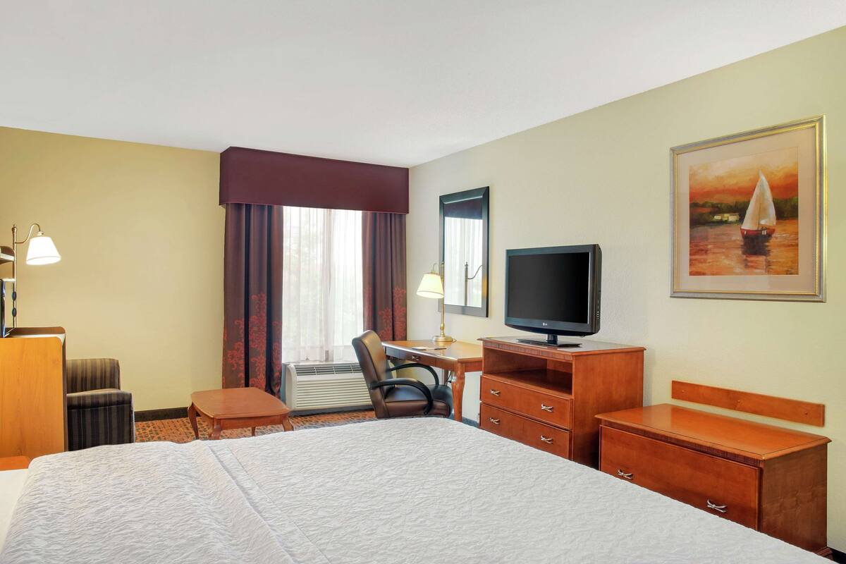 A hotel room with a bed, a desk, a chair, a television, and a painting on the wall.