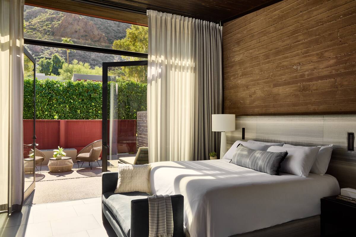A modern bedroom with a large bed, a nightstand, a lamp, and a view of a landscaped outdoor area.