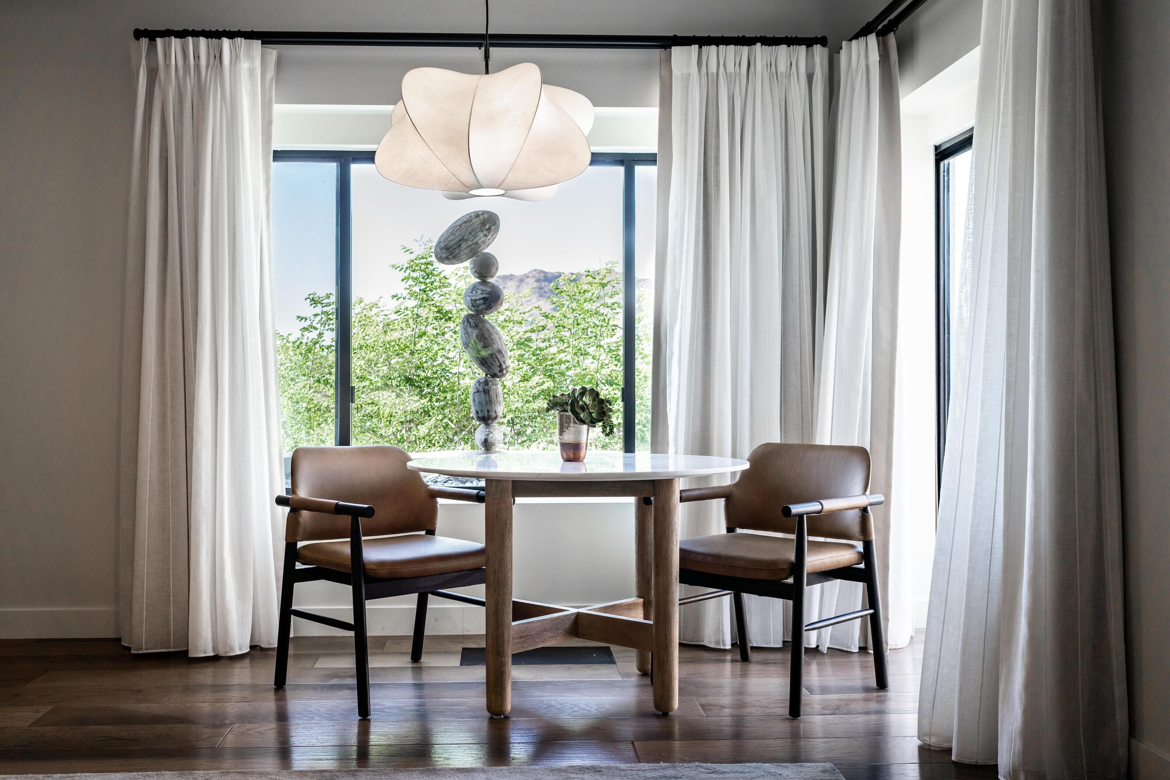 A dining area with a round table, two chairs, a hanging light fixture, and a view of greenery through large windows.