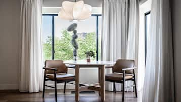 A dining area with a round table, two chairs, a hanging light fixture, and a view of greenery through large windows.