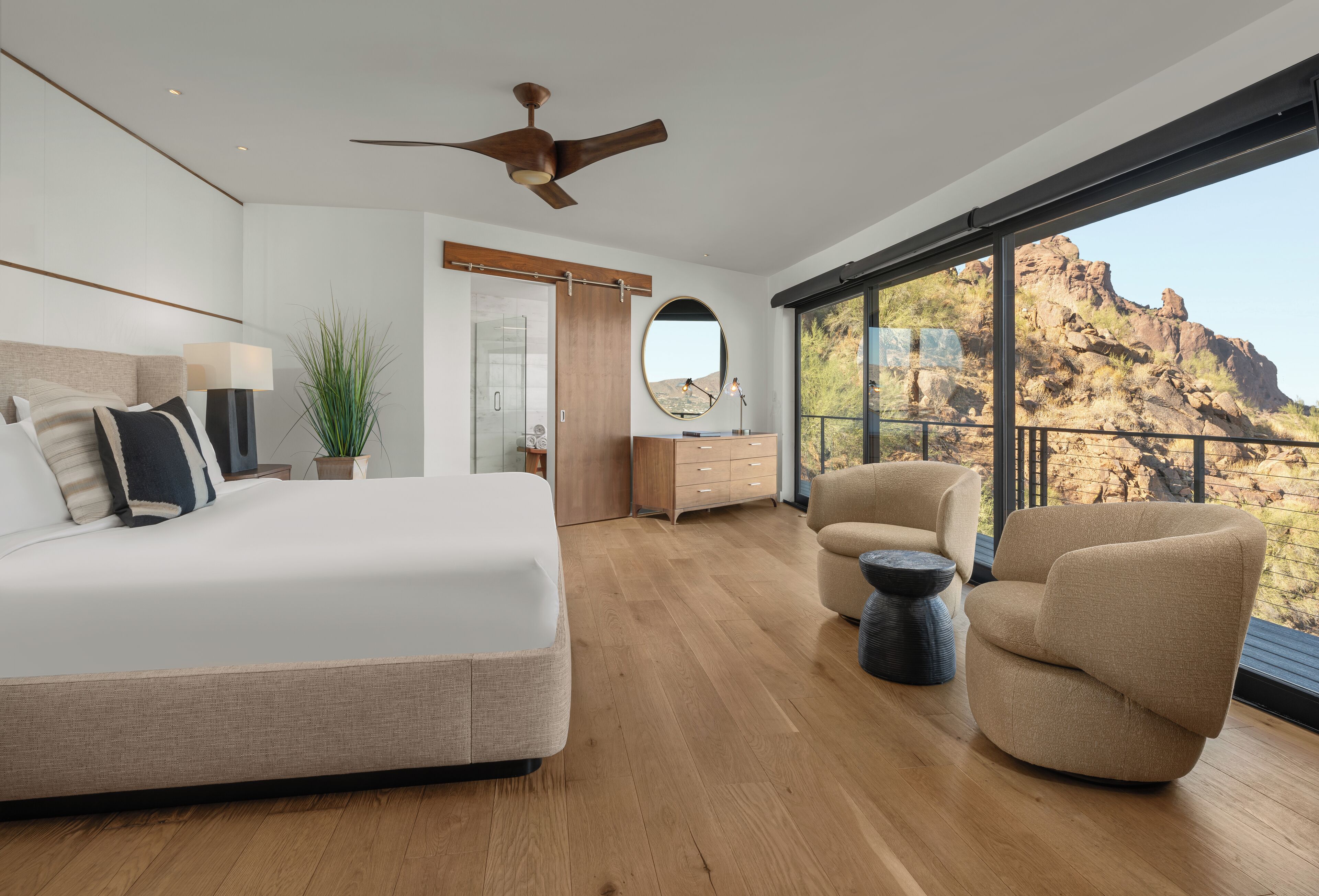 A modern bedroom with a large bed, a ceiling fan, a view of the mountains through large windows, and a balcony with a chair.