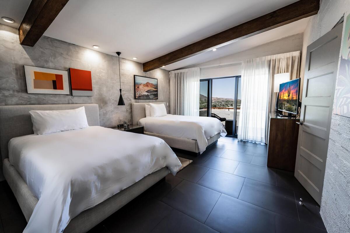 A modern hotel room with two beds, a large window with a view, and artwork on the walls.