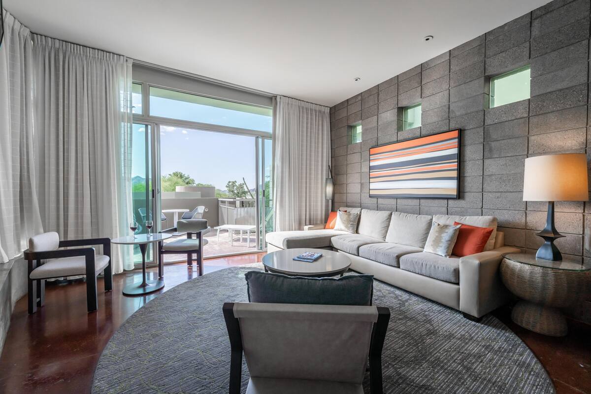 A modern living room with a sofa, armchairs, a coffee table, and a lamp. There is a large window with curtains, a painting on the wall, and a view of the outdoors.