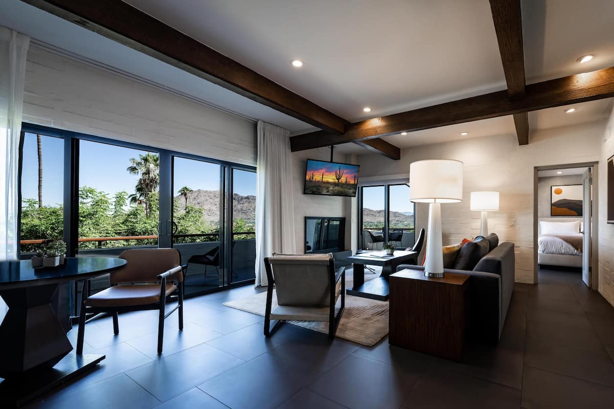 A modern hotel room with a large balcony, a dining area, and a living space with a TV and lamps.