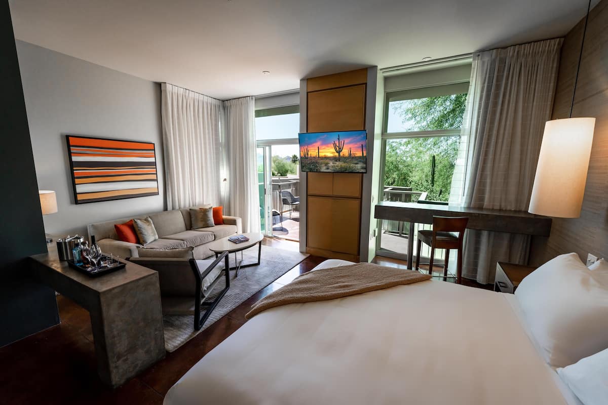 A hotel room with a large bed, a sofa, a small table, and a TV. There is a balcony with a view of the outdoors.