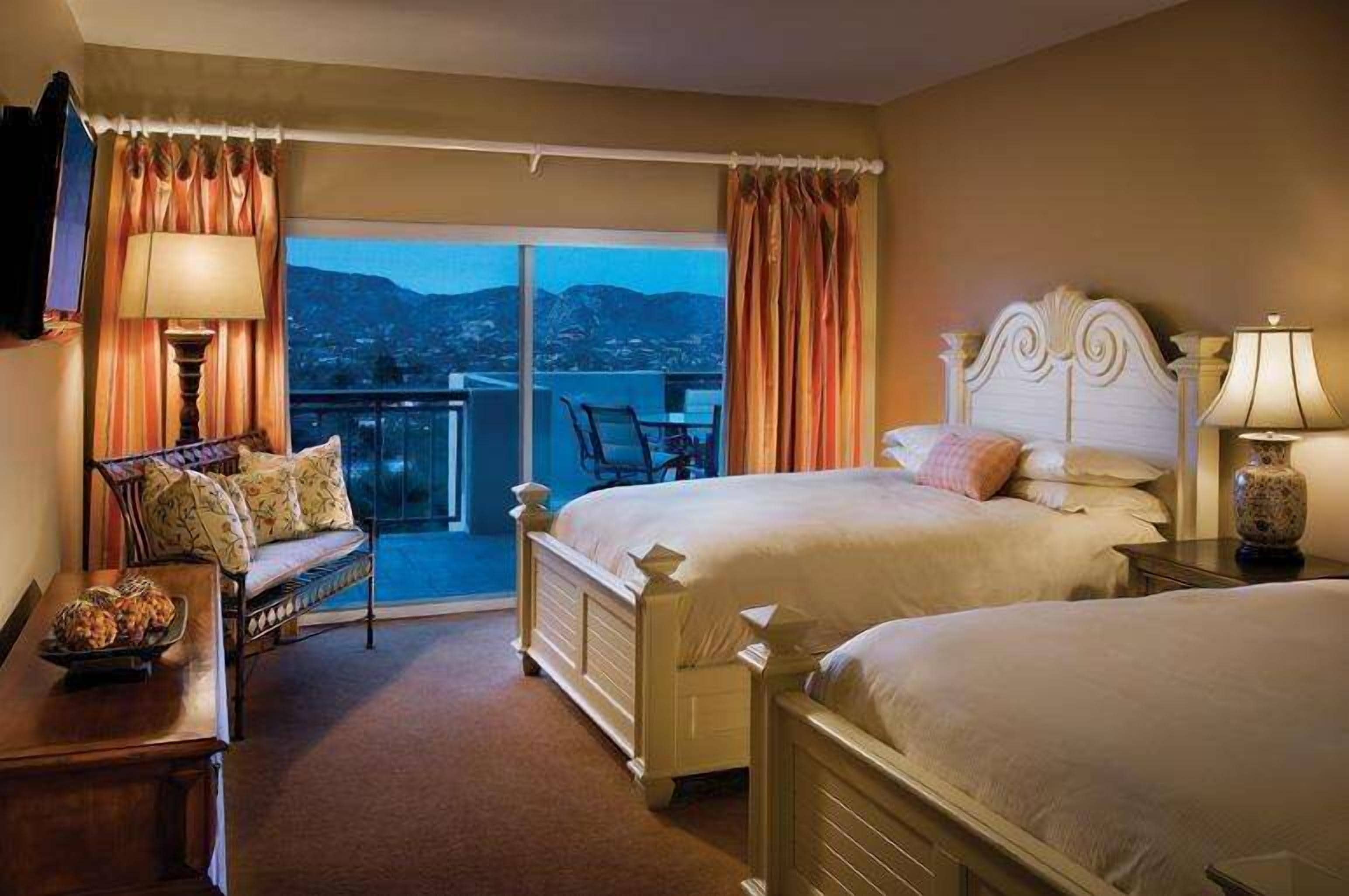 Une chambre d’hôtel avec deux lits, un balcon avec une chaise et une table avec une lampe.