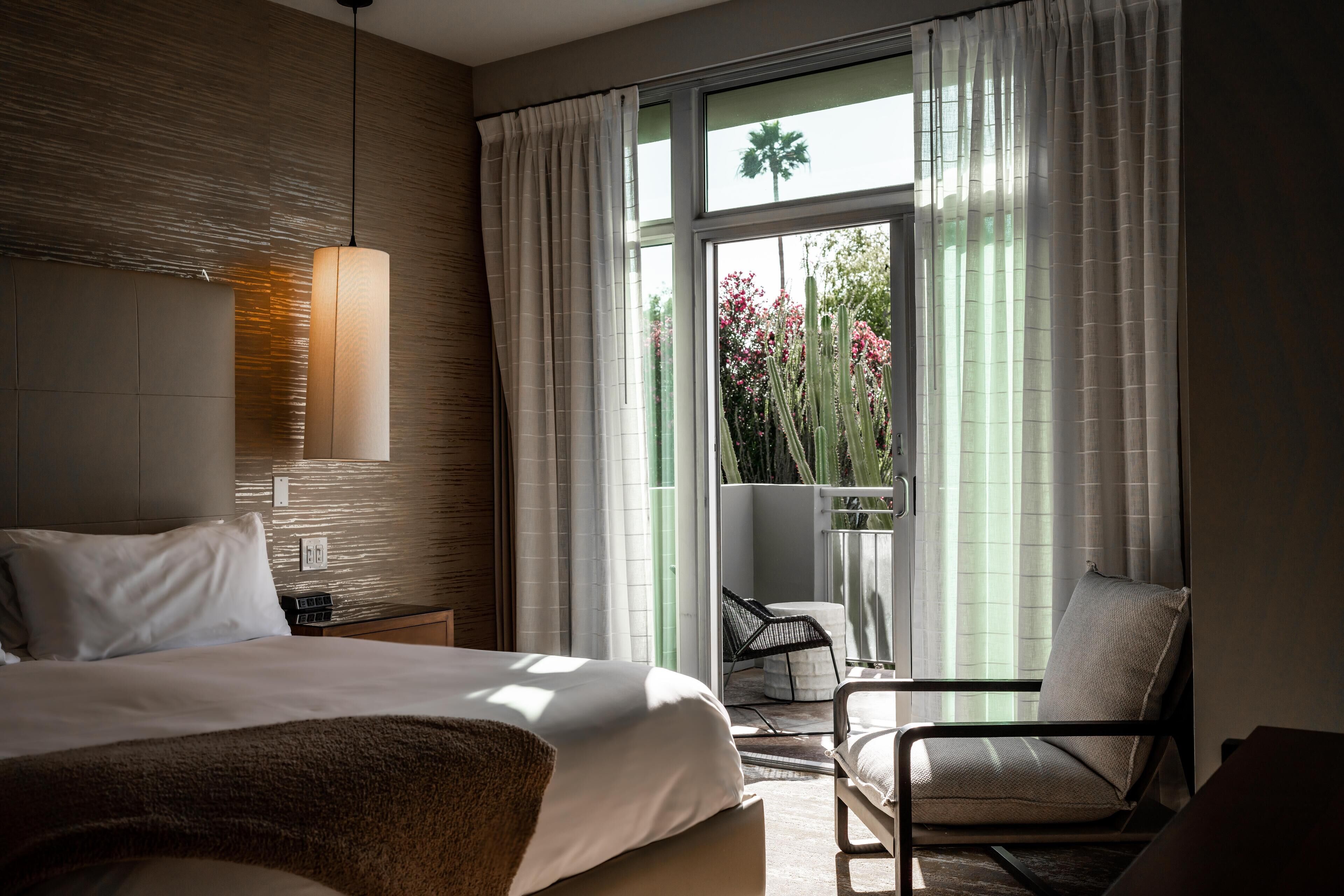 Habitación de hotel con una cama grande, una mesita de noche, una silla y vistas a un balcón con plantas y una palmera.