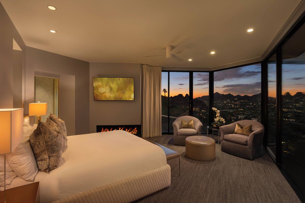 A modern hotel room with a large bed, two armchairs, a small table, and a city view.