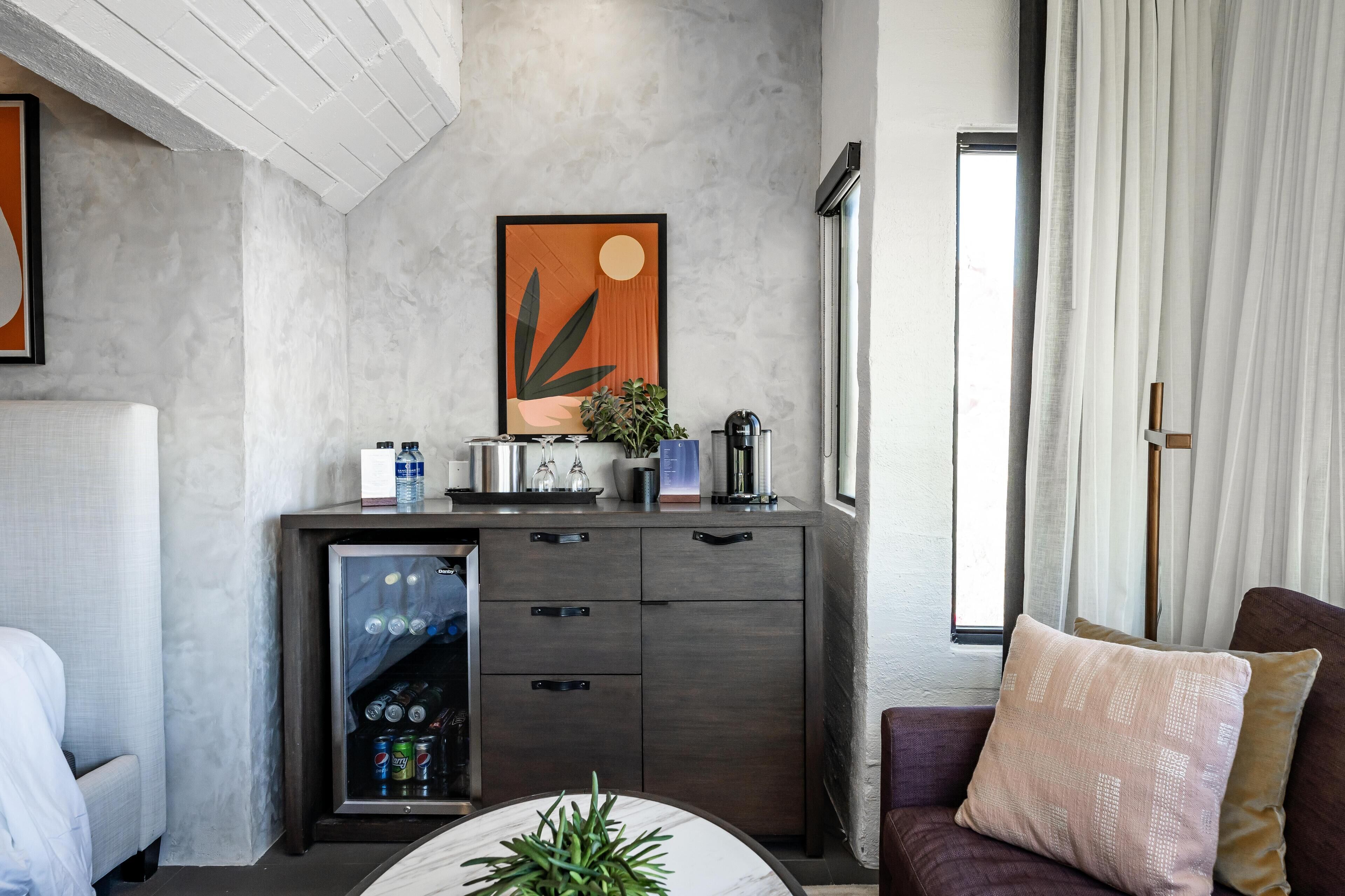 Quarto de hotel moderno com cama, escrivaninha com planta, minibar e uma janela grande com cortinas.