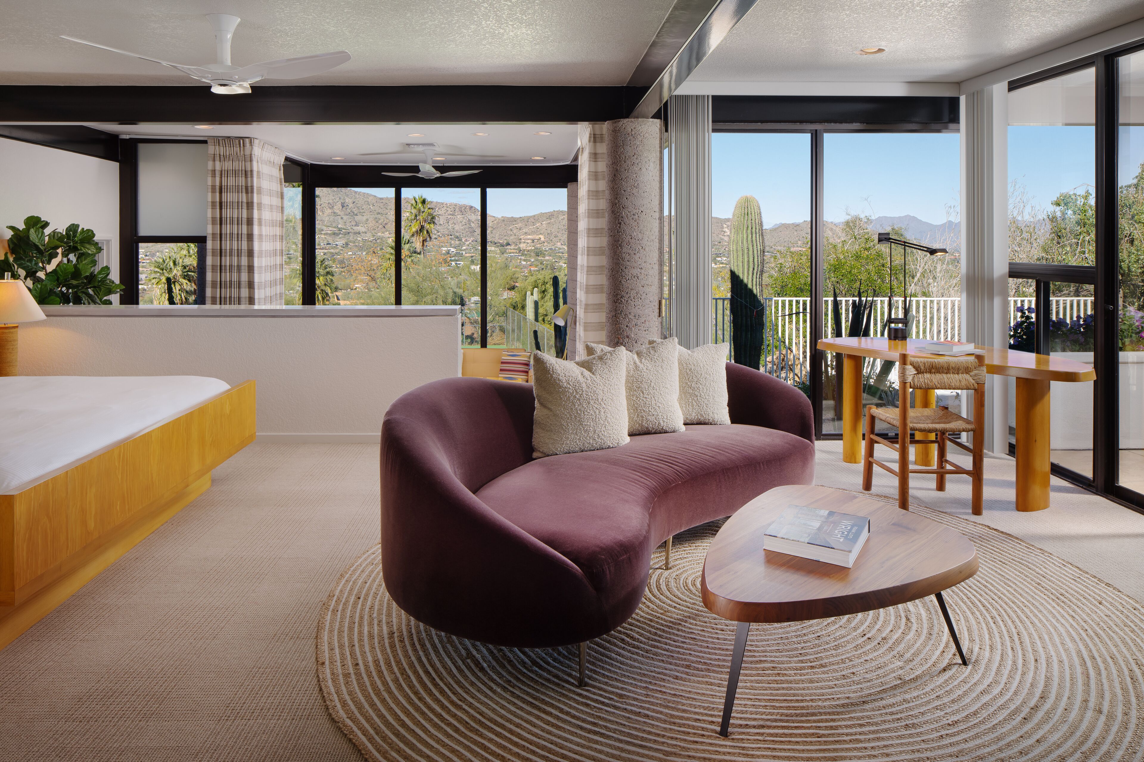 A modern hotel room with a large bed, a sofa, a coffee table, and a balcony with a view of the desert.
